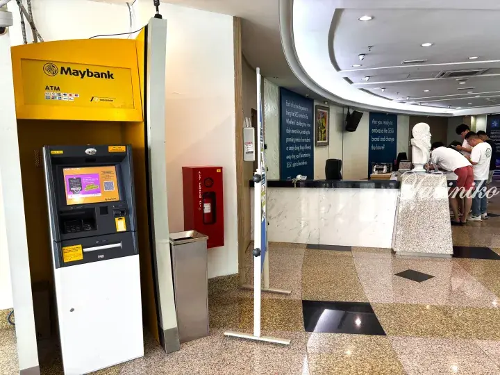 Maybank ATM and fire extinguisher cabinet in a modern building lobby with a reception desk and visitors.