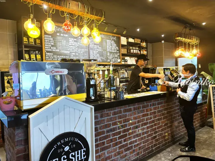 Barista serves customer at He & She Coffee counter with chalkboard menu and La Cimbali espresso machine.
