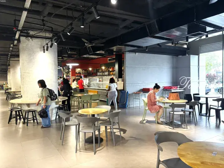 Modern cafe interior with concrete pillars and industrial ceiling, featuring customers dining at round wooden tables near the counter.