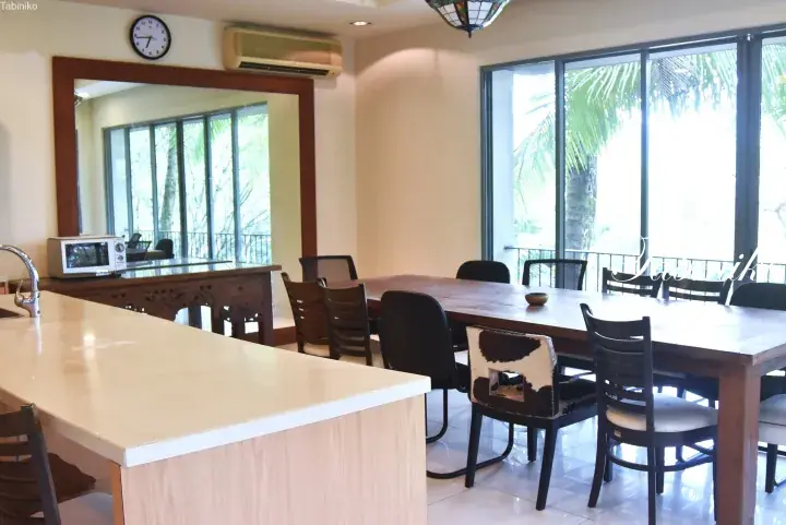 Spacious dining area with large wooden table and mixed chairs, kitchen island, and floor-to-ceiling windows overlooking tropical greenery.