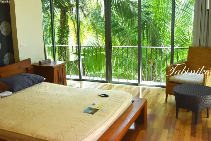 Bright bedroom with wooden furniture and a wicker chair, featuring floor-to-ceiling glass doors opening to a lush tropical garden view.