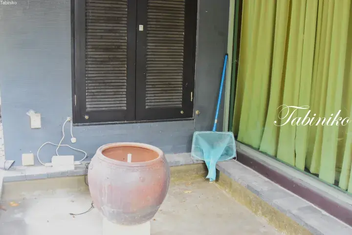 Terracotta water jar and blue net on a balcony with green curtains and louvered windows.