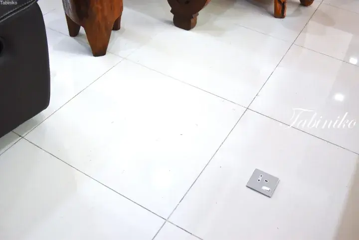 Close-up of a modern floor socket on white tiled flooring in a property, with wooden furniture legs visible.