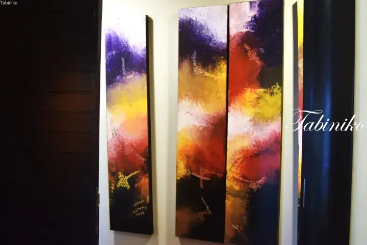 Three tall abstract paintings with purple, yellow, and red hues hang on a white wall.