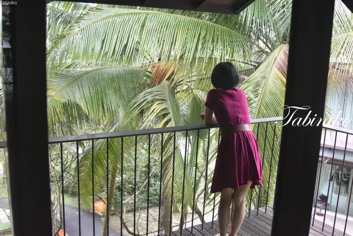 Woman in a burgundy dress standing on a wooden balcony overlooking lush tropical palm trees at a resort.