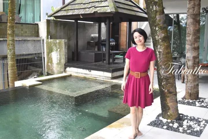 Woman in a pink dress standing barefoot by a private pool at a tropical villa with a wooden gazebo.