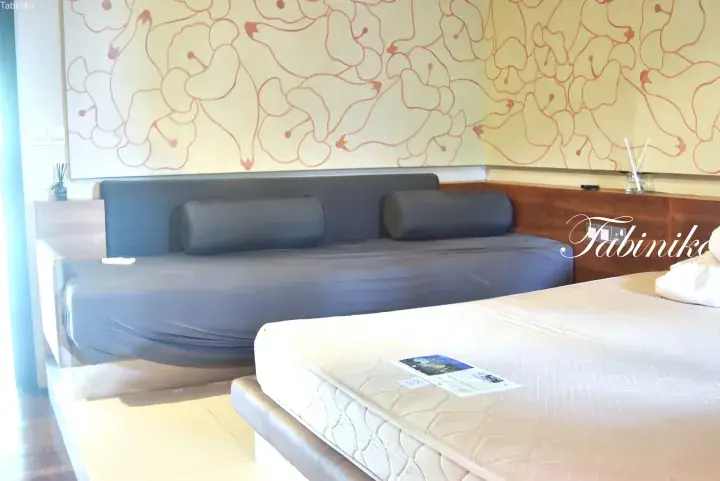 Modern hotel room with a grey sofa bed and a double bed featuring a patterned mattress against a pear-themed wall.