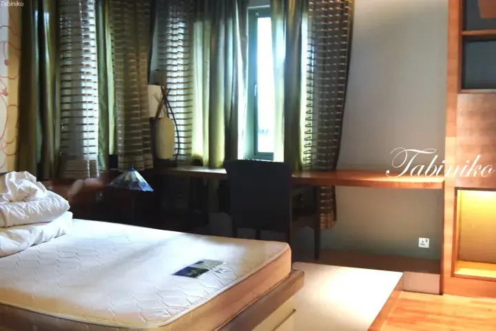 Hotel room interior featuring a bed with white linens, a wooden desk, and green curtains.