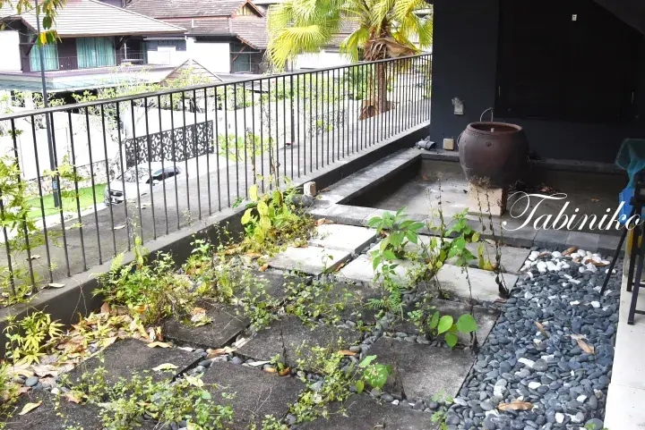 Overgrown garden path with stone slabs and pebbles at a property, with a large ceramic pot and balcony railing visible.