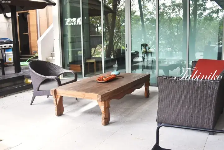 Rustic wooden coffee table with wicker chairs on a tiled patio outside a glass-walled villa.