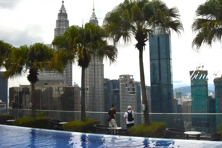 View from a rooftop pool featuring palm trees and the iconic Petronas Twin Towers in the Kuala Lumpur skyline.