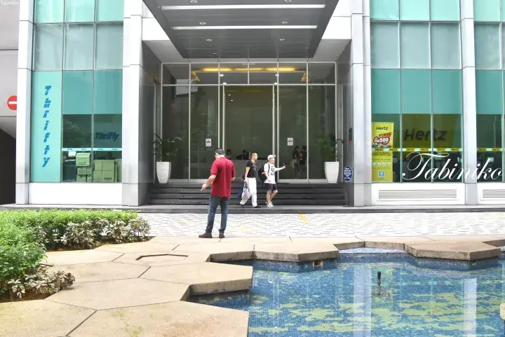 Modern building entrance with glass facade, Thrifty and Hertz car rental signs, and a decorative water feature in the foreground.