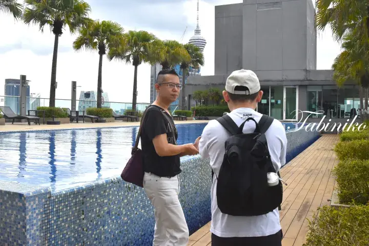 Two men conversing by a rooftop pool with Kuala Lumpur Tower visible in the background.