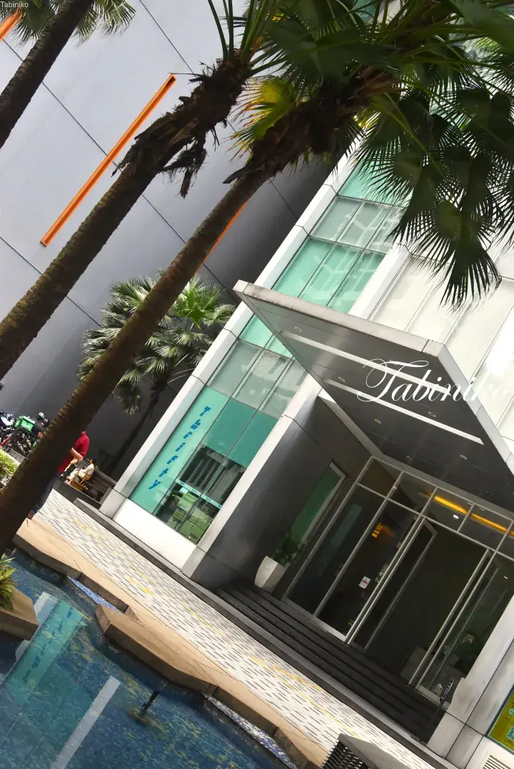 Modern building entrance with glass facade, palm trees, and a decorative pool.