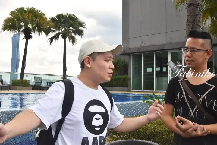 Two men conversing by a rooftop pool with palm trees and a modern skyscraper in the background.