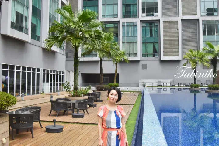 Woman standing by a swimming pool deck at a modern residential complex with palm trees and lounge chairs.