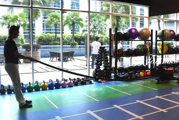 Man using battle ropes in a modern gym overlooking a pool and palm trees.