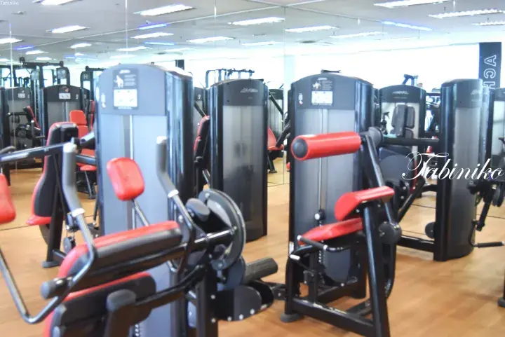 Rows of modern weight training machines with red and black upholstery in a well-lit gym with mirrored walls