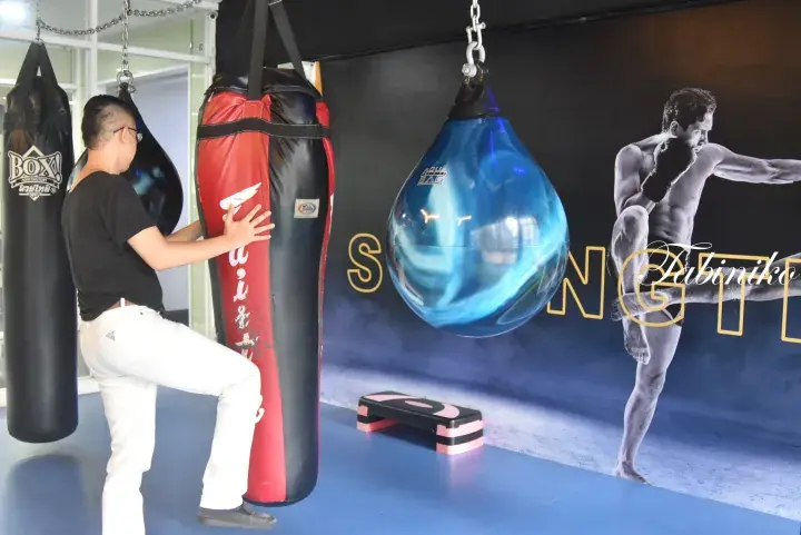 Man in black shirt and white pants practicing Muay Thai kicks on a heavy bag at a gym with boxing equipment.