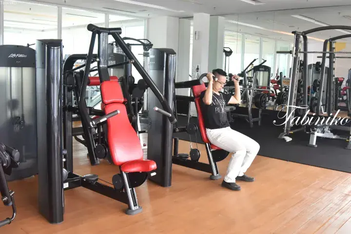 Man exercising on a shoulder press machine in a well-lit gym with wooden floors and mirrors.
