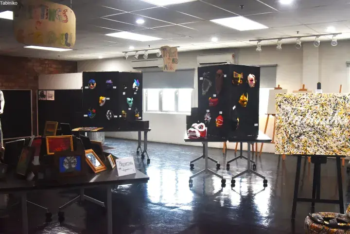 Art exhibition featuring colorful masks on black panels, framed paintings, and abstract canvas art in a gallery space.