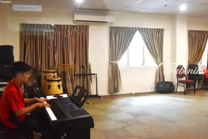 Musician plays keyboard in a bright practice room with conga drums and soundproofing curtains.