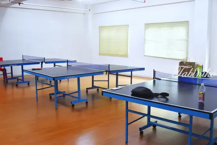 Multiple blue Nittaku table tennis tables arranged in a bright indoor sports hall with wooden flooring.