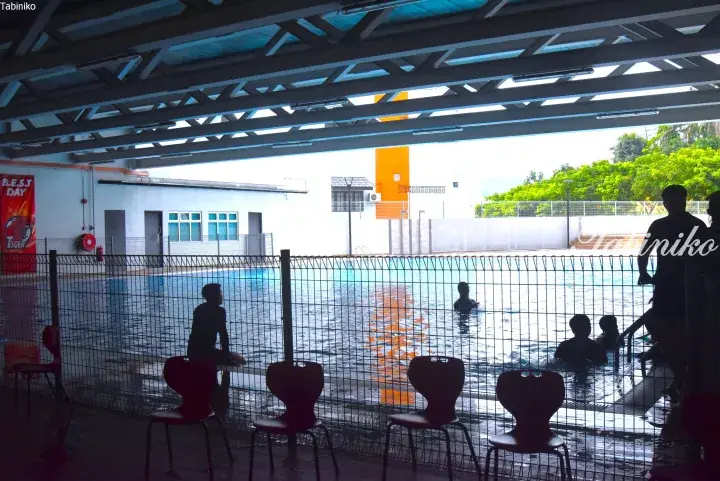 People swimming in an outdoor pool viewed from under a shaded structure with red chairs.