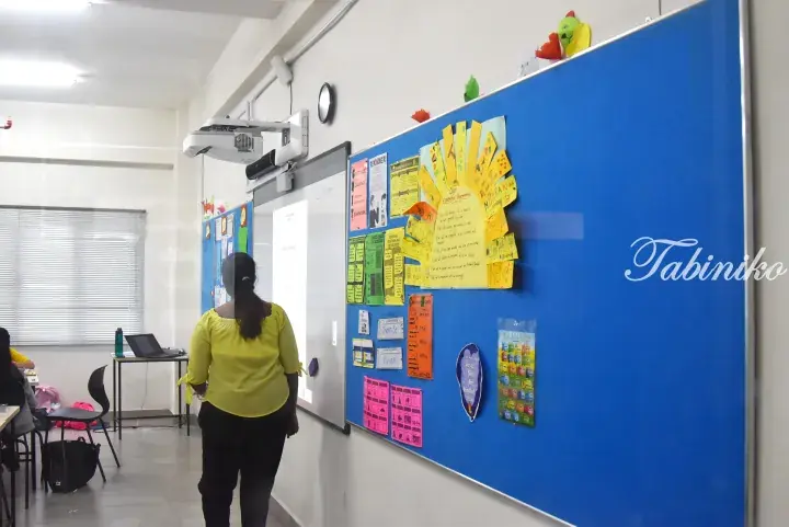 Teacher in yellow shirt facing a blue bulletin board with student artwork and a whiteboard in a bright classroom.