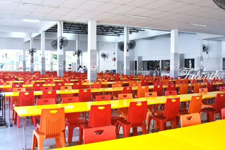 Spacious school cafeteria with rows of yellow tables and red plastic chairs under ceiling fans.