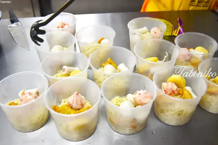 Multiple plastic cups filled with instant noodles, fish balls, and tofu being prepared for a meal.