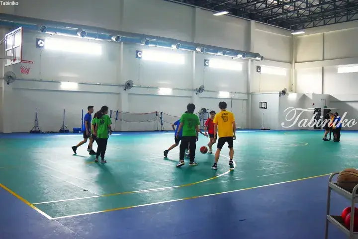 Students in colorful shirts play basketball on a green court inside a spacious indoor sports hall with blue flooring.