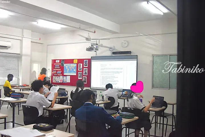Students in a modern classroom using laptops and reviewing notes while a presentation is displayed on a large screen.