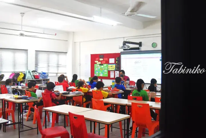 Students in a bright classroom with orange chairs and a teacher at the front near a projector screen.