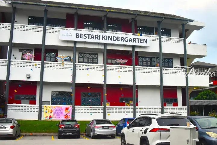 Exterior of Bestari Kindergarten building with white and red facade, balconies, and parked cars in front.