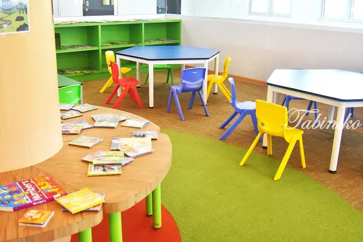 Colorful children’s chairs and tables in a bright kindergarten reading corner with books and green shelving.