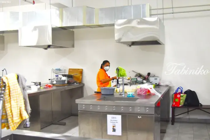 Woman in orange shirt and mask preparing food in a commercial kitchen with stainless steel counters and ventilation hoods.