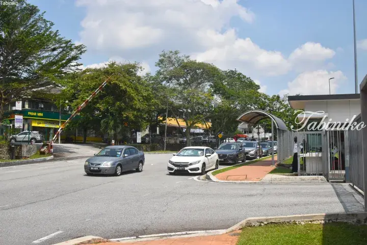 Cars waiting at a security checkpoint entrance in Malaysia, with a guard booth and a mini market visible nearby.