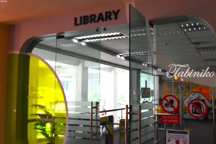 Glass entrance to a modern library with a yellow accent wall and signs prohibiting food and bags.