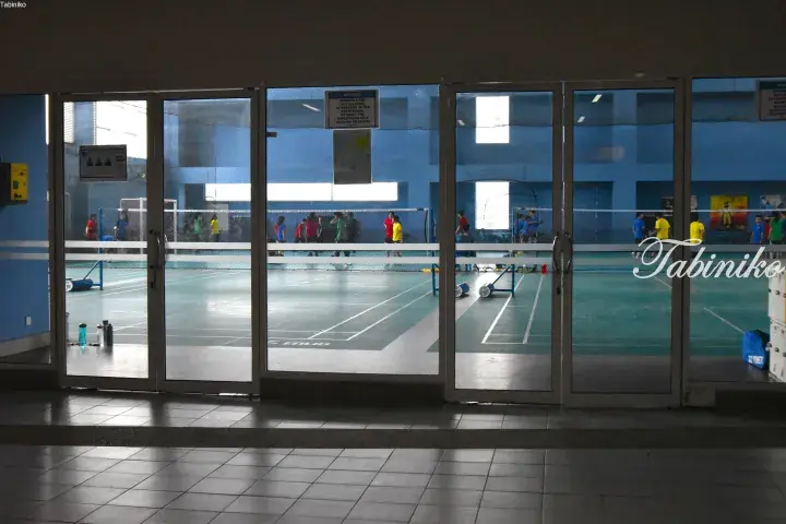 View through glass doors into a school sports hall with students playing badminton on green courts.