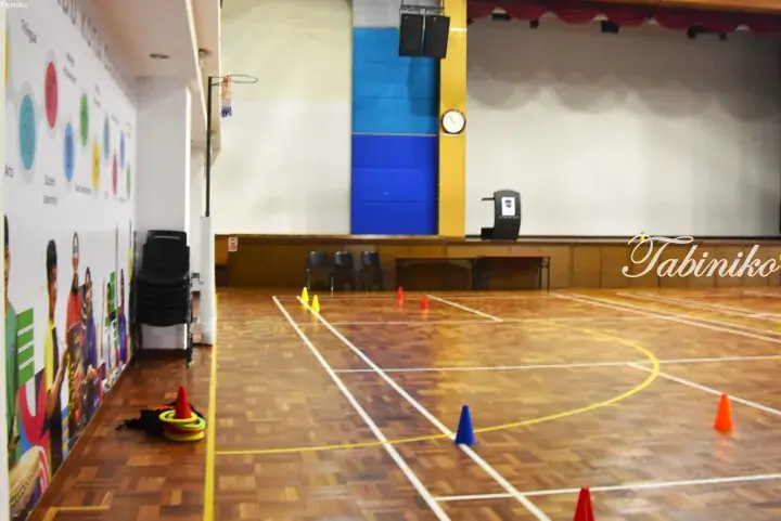 Indoor gymnasium with wooden flooring, sports cones, and a stage area in a Malaysian school or community center.