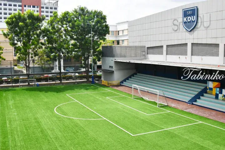Green artificial turf football pitch with white goalposts and blue stadium seating at KDU College in Malaysia.