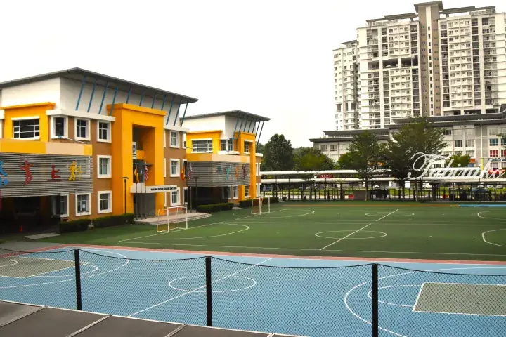 Colorful modern school building with yellow accents, green sports field, and high-rise apartments in the background.