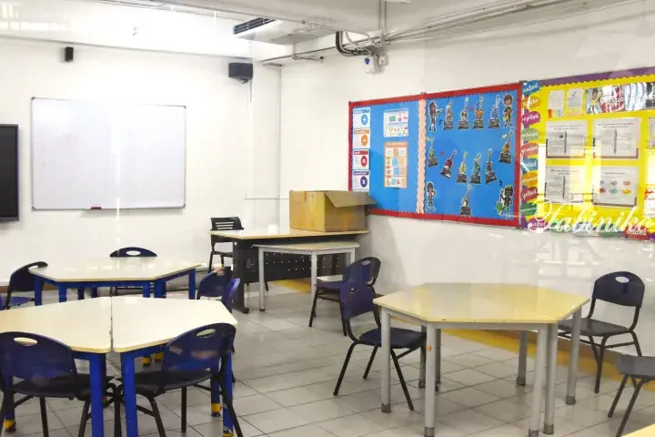 Bright classroom with blue chairs, wooden tables, whiteboard, and colorful educational posters on the wall.