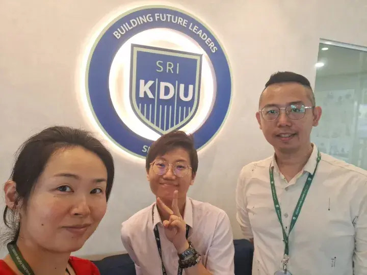 Three people pose in front of the Sri KDU logo with the motto Building Future Leaders.