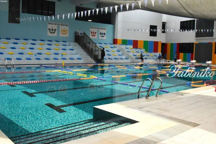 Indoor swimming pool with blue and yellow spectator seating, lane ropes, and colorful wall accents.