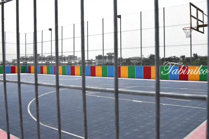 View of a blue basketball court with colorful striped walls, seen through a metal fence.