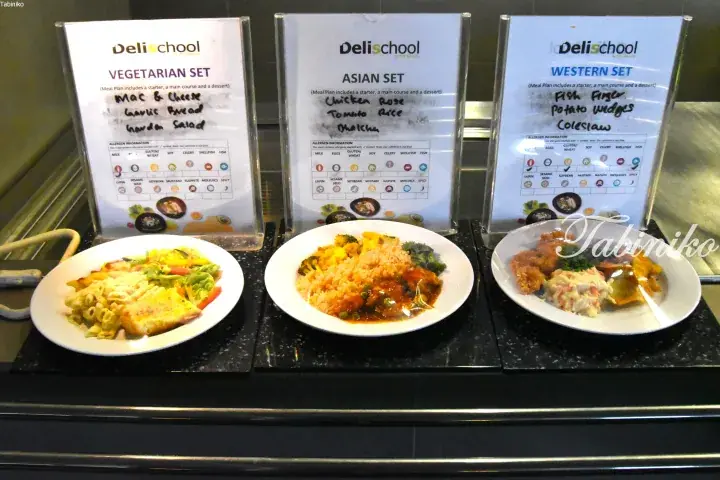 Three Delischool cafeteria meal sets displayed with signs: Vegetarian Mac & Cheese, Asian Chicken Rose, and Western Fish Finger.