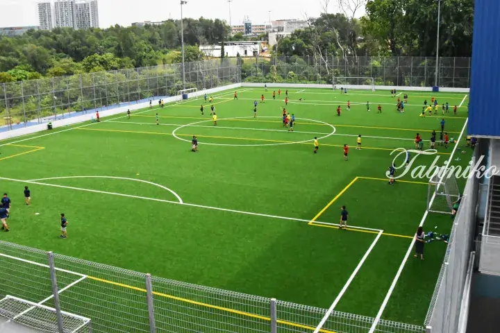 Aerial view of a vibrant green artificial turf football pitch with players in action, surrounded by high fences and lush trees in Malaysia.