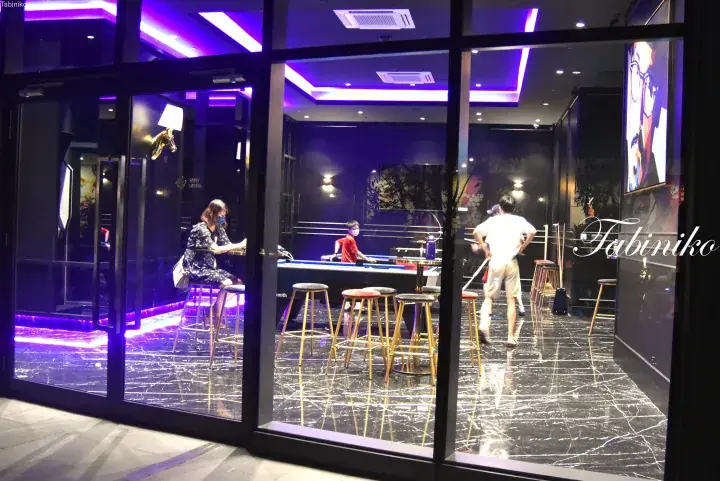 Modern lounge with purple lighting, black marble floors, and patrons playing pool behind glass windows.